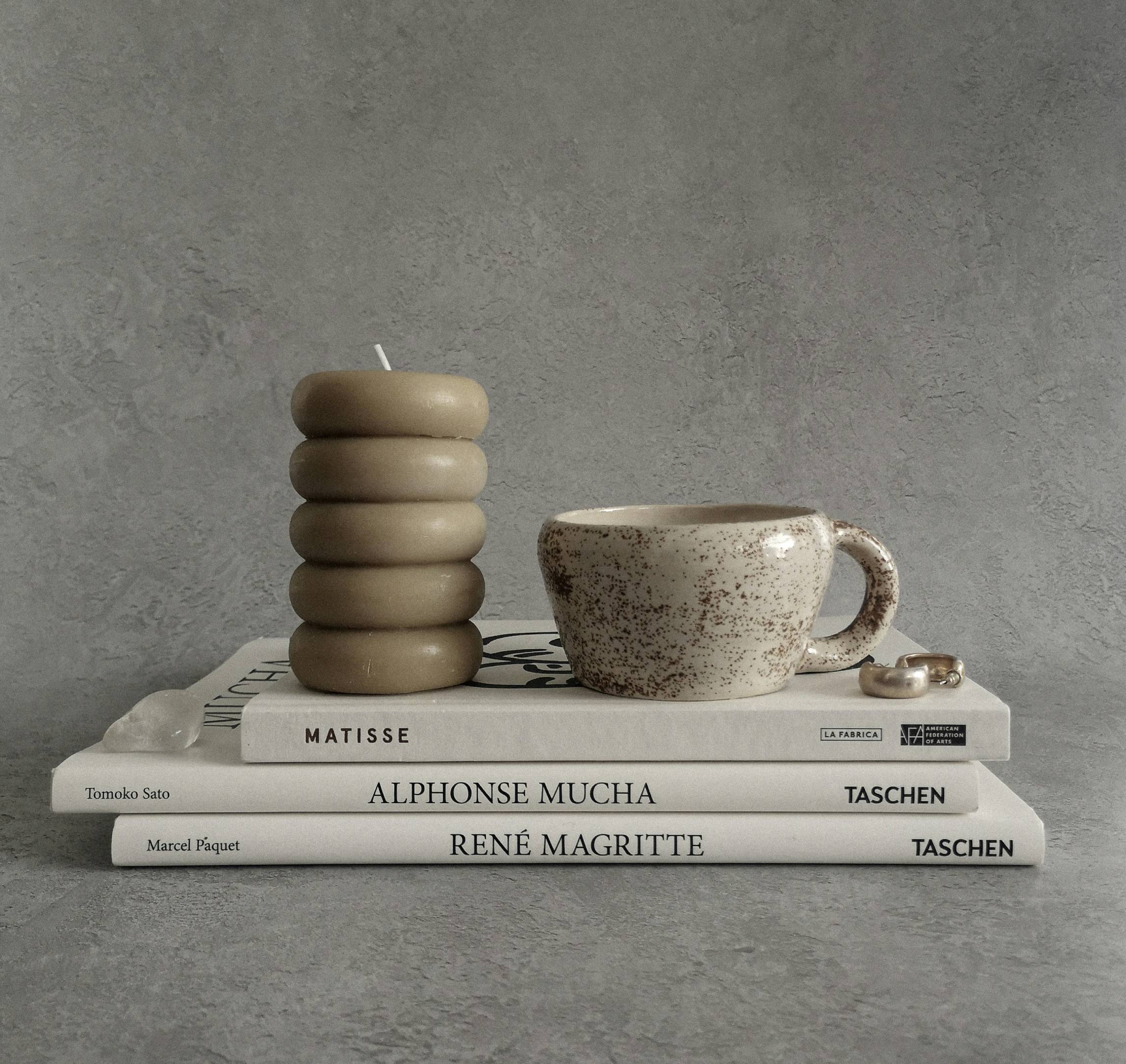 Minimalist home decor flat lay featuring a stacked beige pillar candle, a speckled ceramic coffee cup, and silver rings styled on art books about Matisse, Alphonse Mucha, and René Magritte – modern aesthetic home accessories from Colliam.